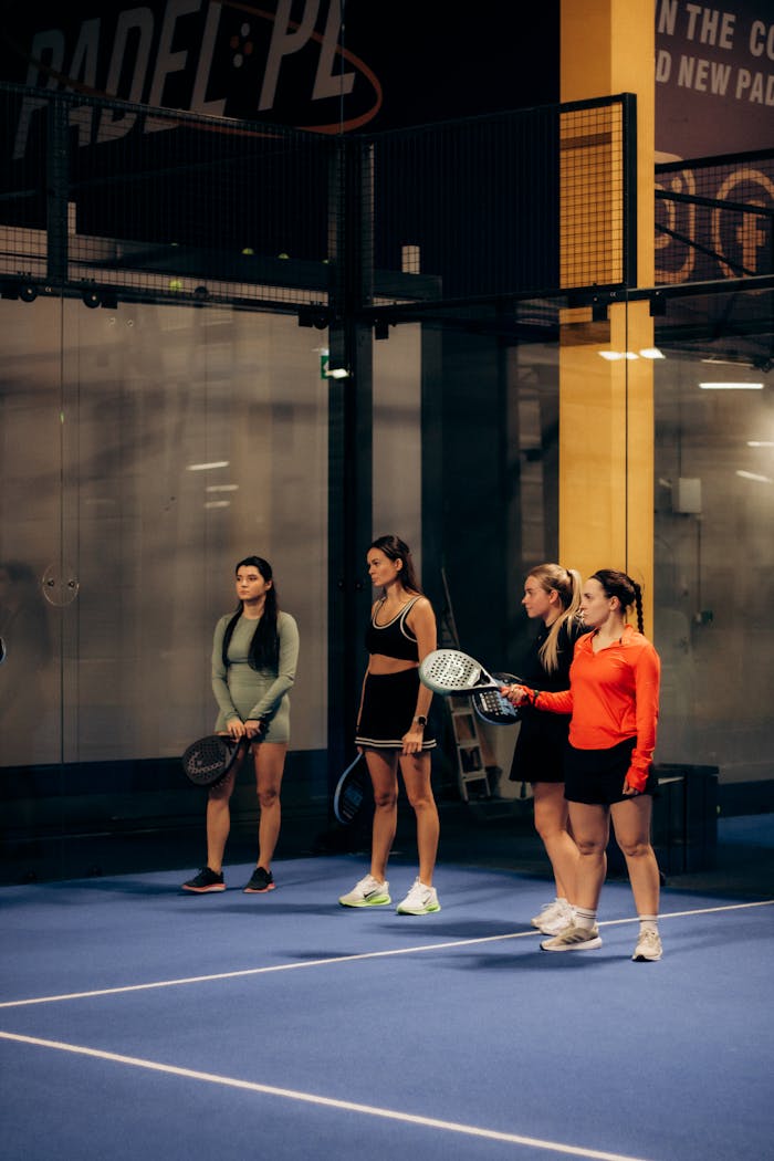 Four women holding rackets stand on a blue padel court, ready to play indoors.